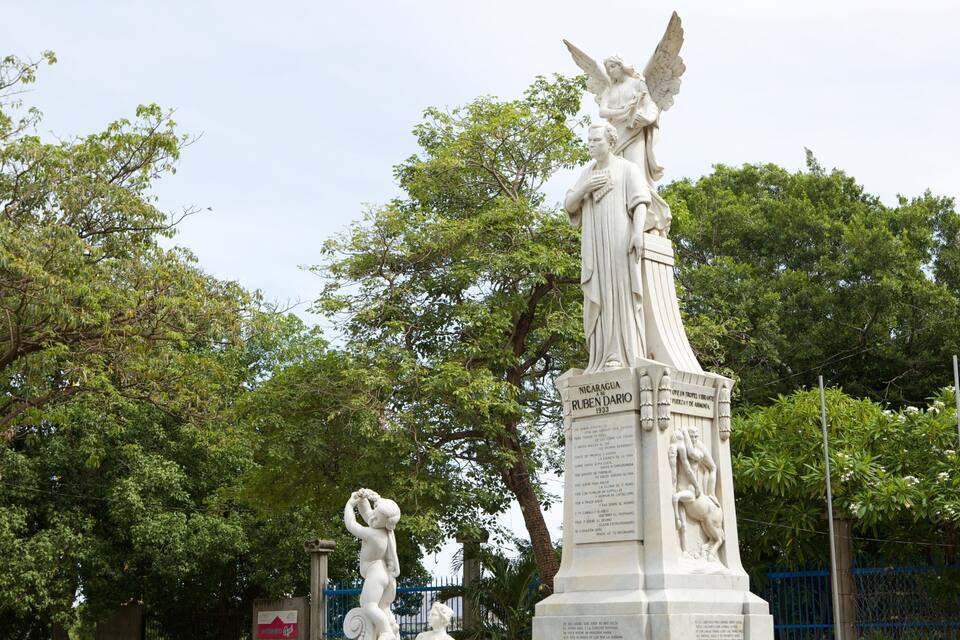 Una estatua de una mujer sosteniendo una lira, rodeada de árboles y una valla.