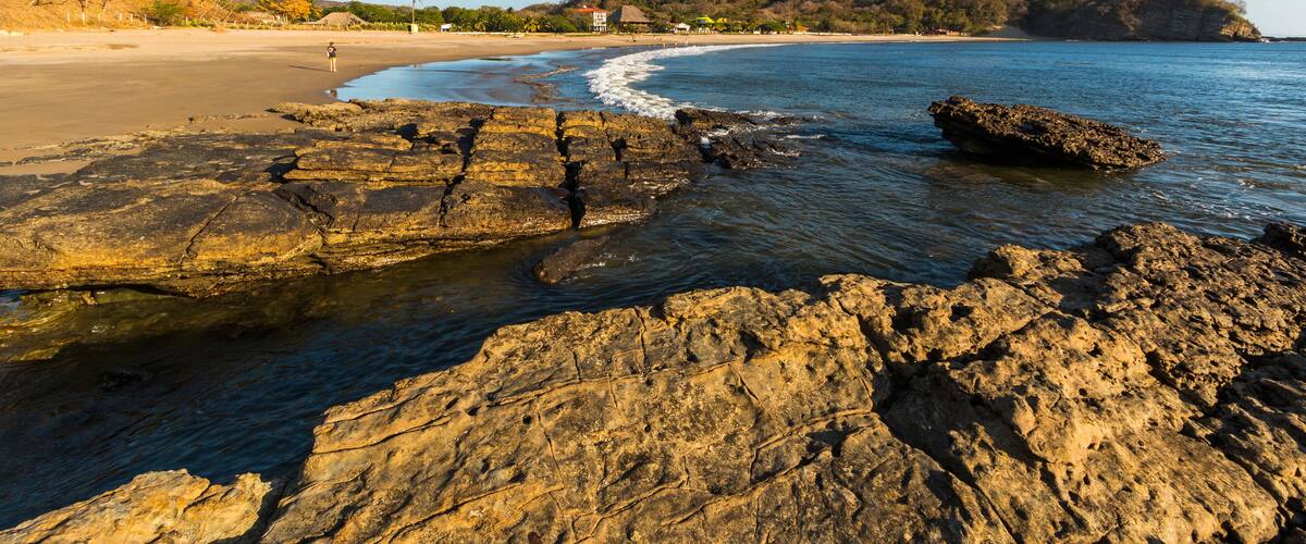 A rocky shoreline with clear water, a sandy beach, and a distant hill with trees.