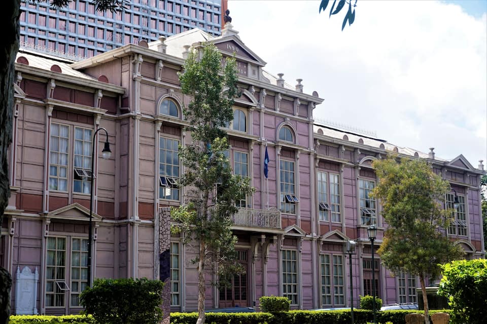 A historic building with arched windows and a balcony, surrounded by well-maintained hedges.