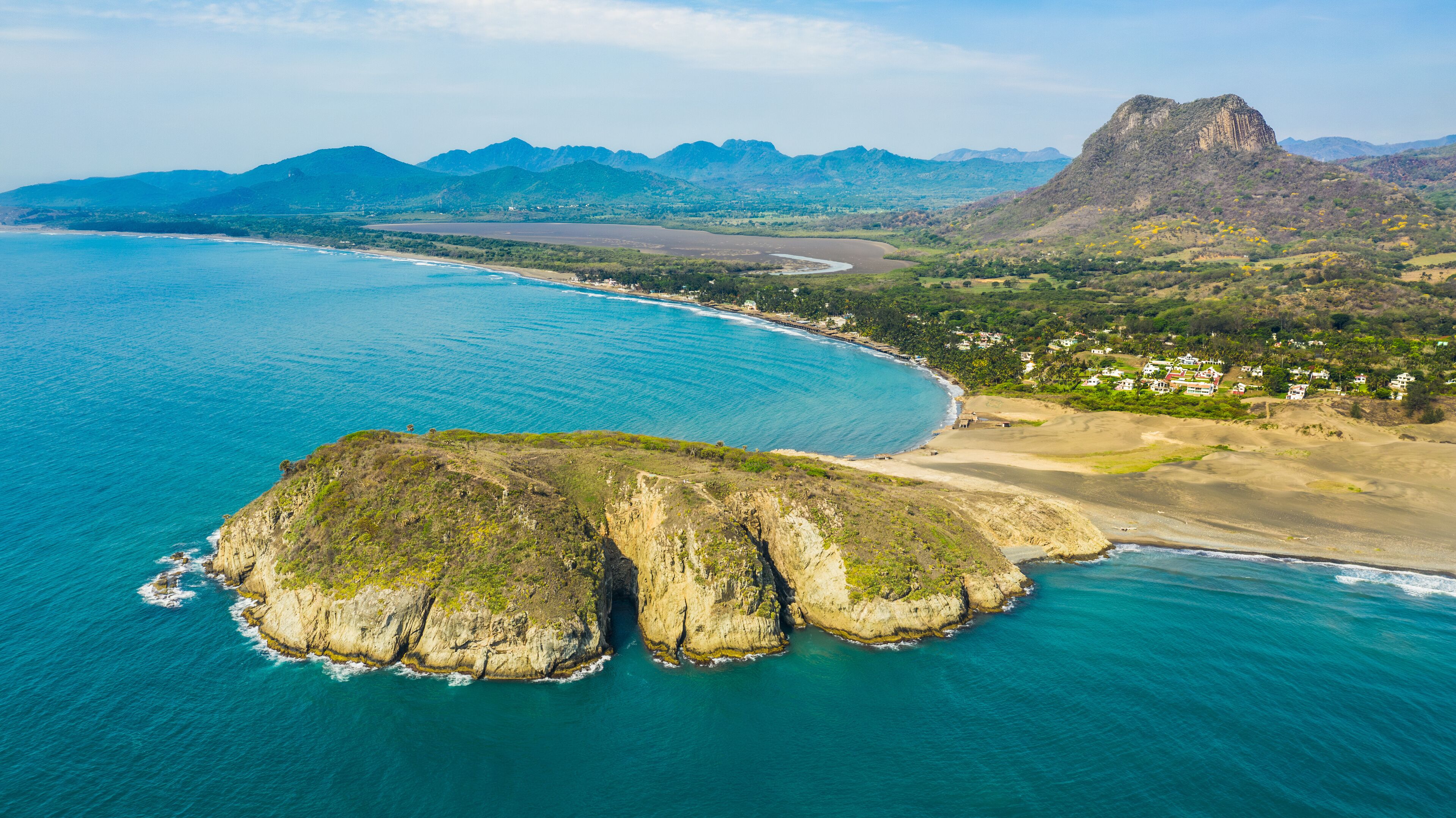 A coastal landscape with a prominent rock formation, a beach, and distant mountains.