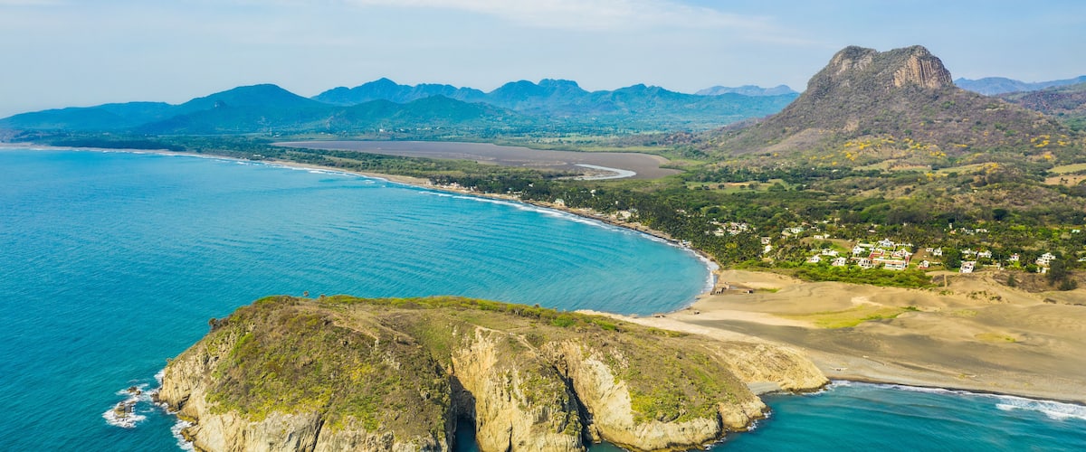 A coastal landscape with a prominent rock formation, a beach, and distant mountains.