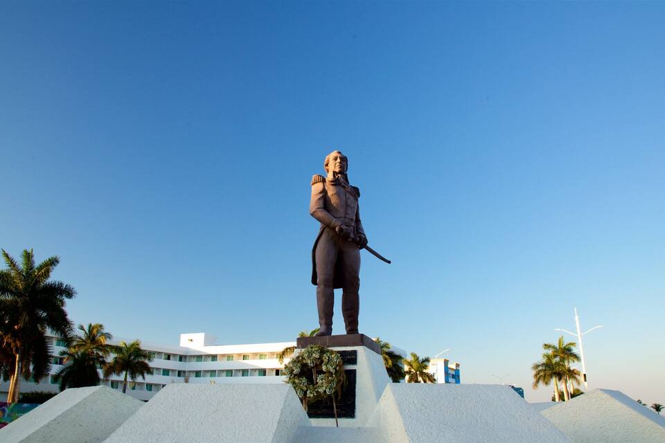Una estàtua d'un militar damunt un pedestal, amb palmeres i un edifici al fons.