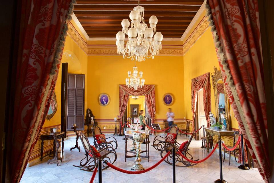 Une pièce richement décorée, avec un lustre, des rideaux rouges et une barrière délimitée par une corde rouge.