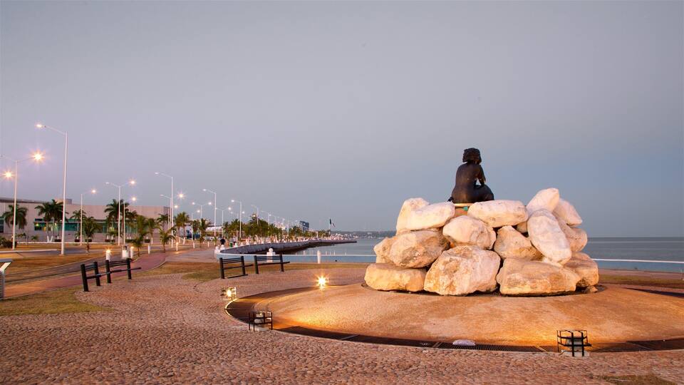Escultura d'una figura asseguda sobre un munt de roques, envoltada de bancs i fanals, amb vista a una platja.