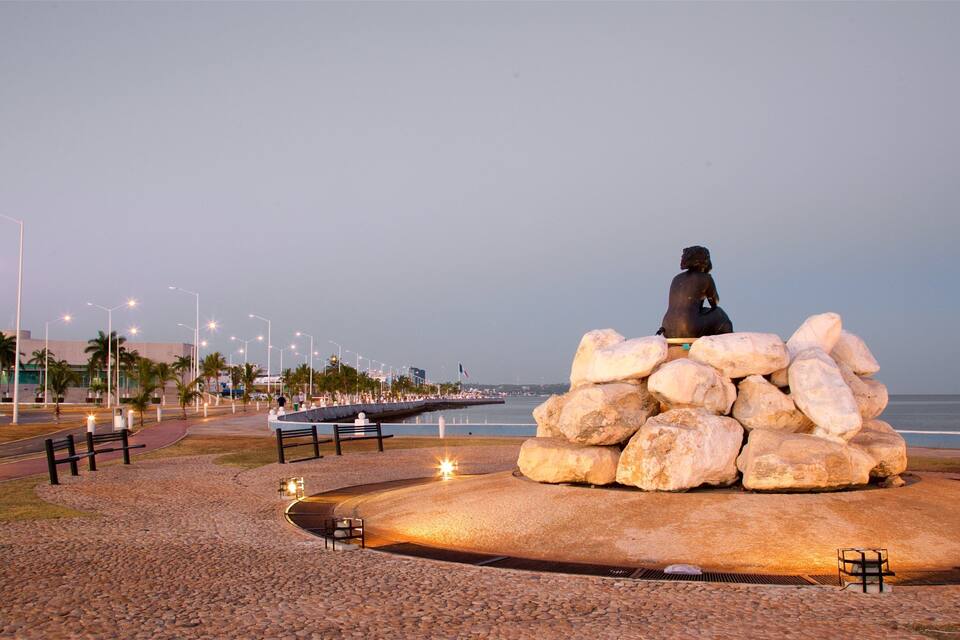 Una escultura de una figura sentada sobre un montón de rocas, rodeada de bancos y farolas, con vista a una playa.