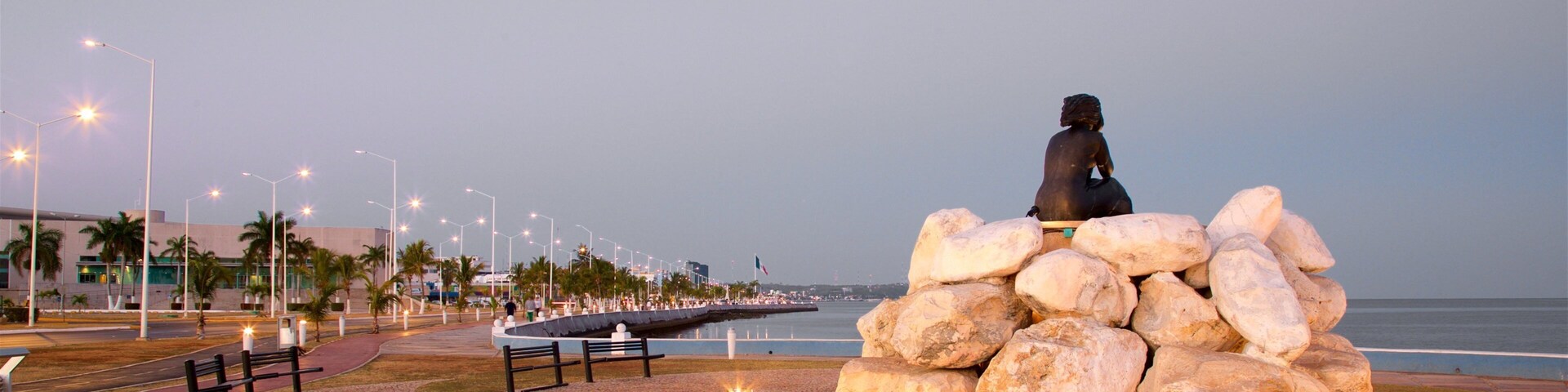 Una escultura de una figura sentada sobre un montón de rocas, rodeada de bancos y farolas, con vista a una playa.