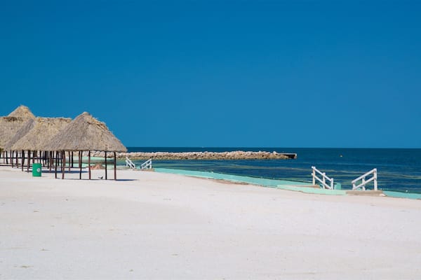 Ein Strand mit strohgedeckten Hütten, einem felsigen Wellenbrecher und blauem Himmel.