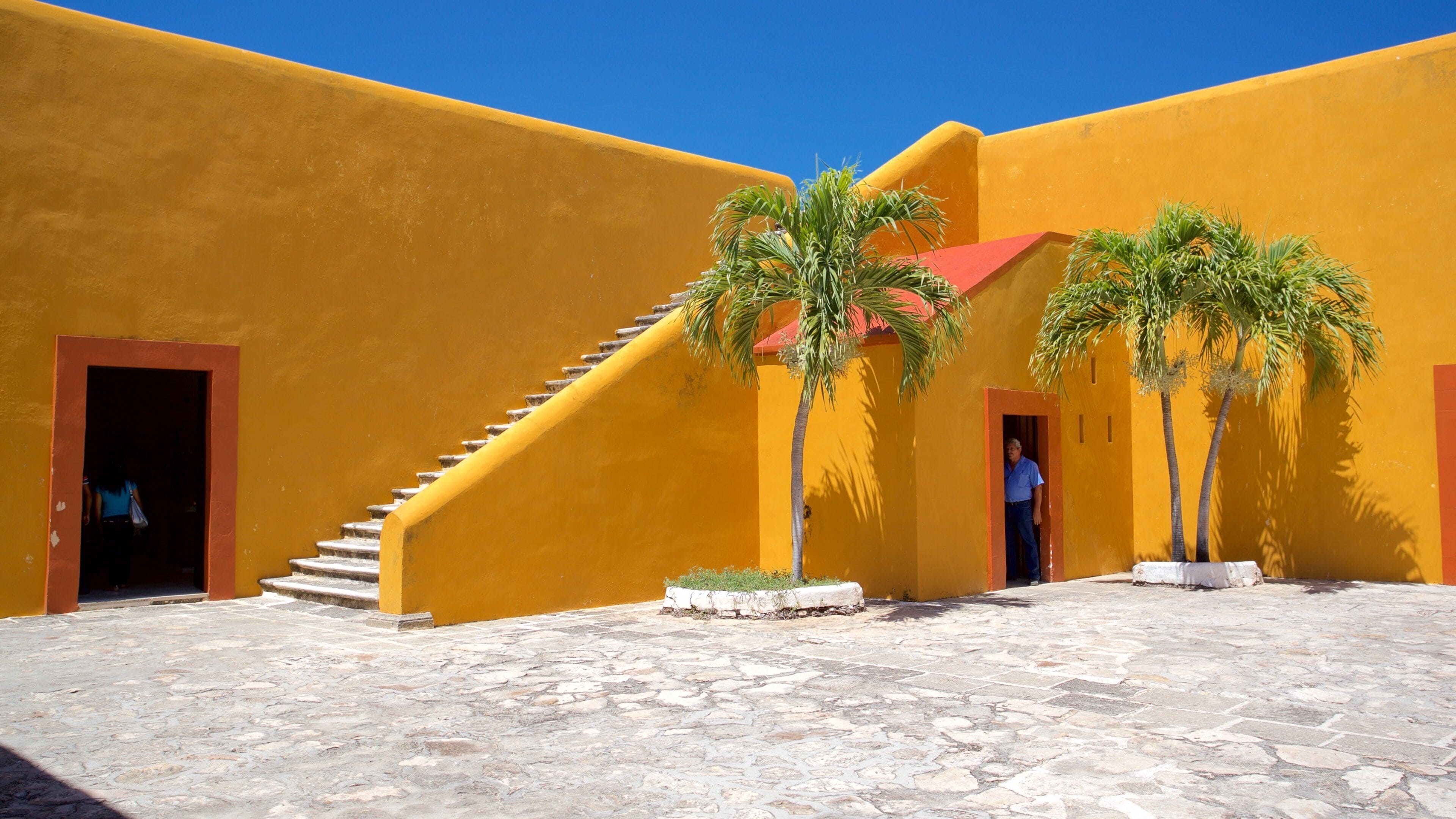 A courtyard with yellow walls, a stone floor, and palm trees.