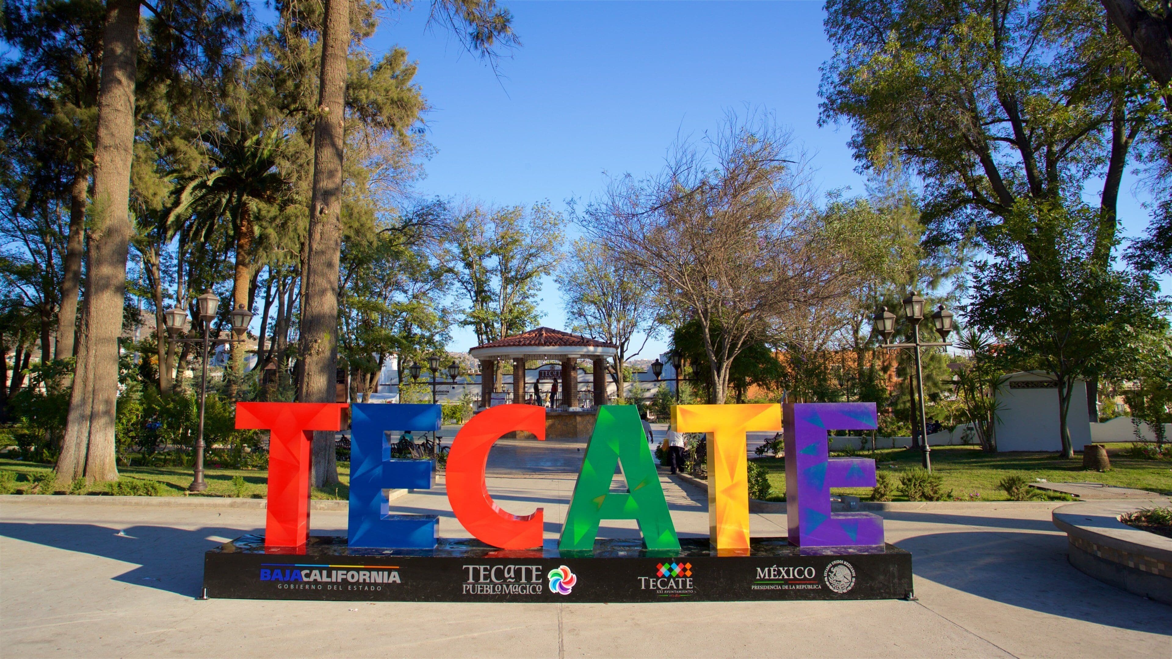 Un letrero grande al aire libre con la palabra "TECATE" en letras coloridas, rodeado de árboles y un pabellón al fondo.
