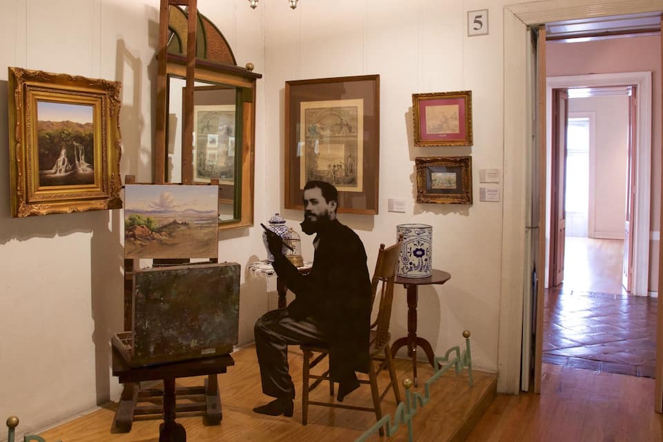 Een galerij met tentoongestelde schilderijen, een houten vloer en een deur naar een andere ruimte.