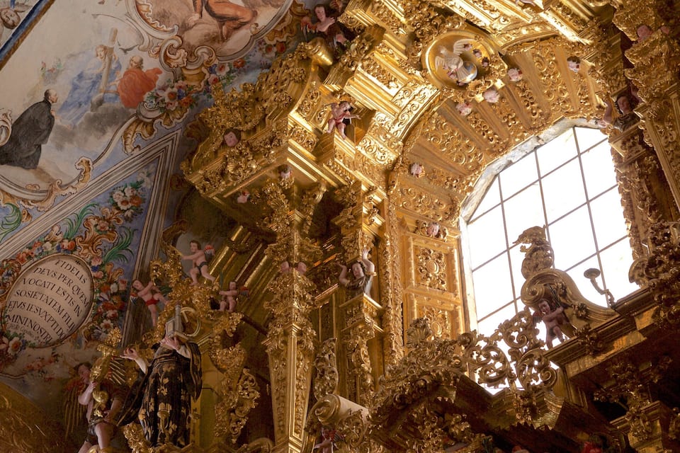 Interior ornamentado com detalhes dourados, querubins e uma janela grande.