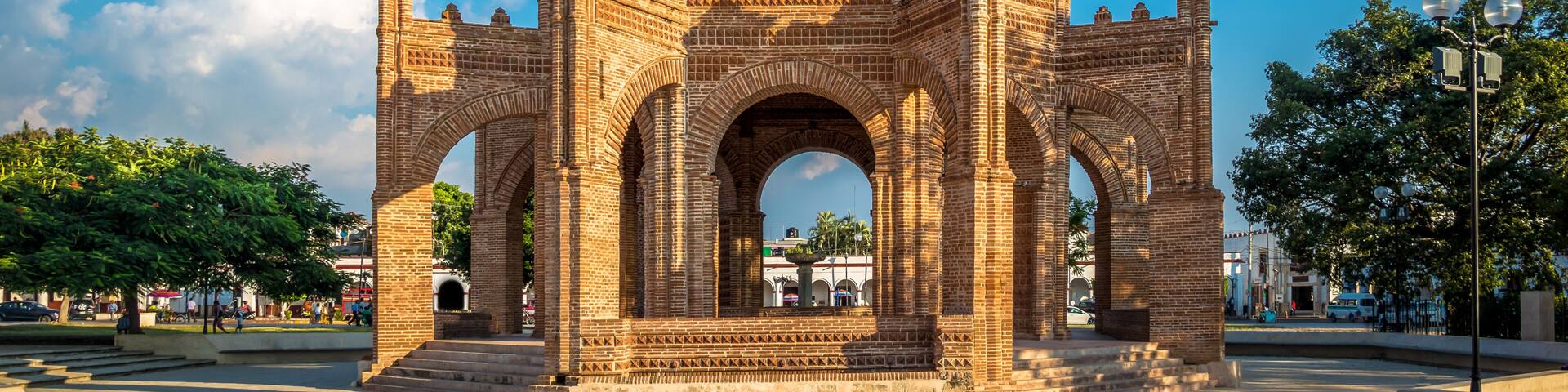 A large, ornate brick archway with multiple arches and a central dome, set in a public square with trees and a clear blue sky.
