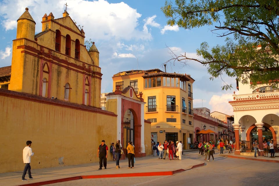 Uma praça histórica com arquitetura colonial, incluindo uma igreja com um campanário e um edifício com um arco vermelho.
