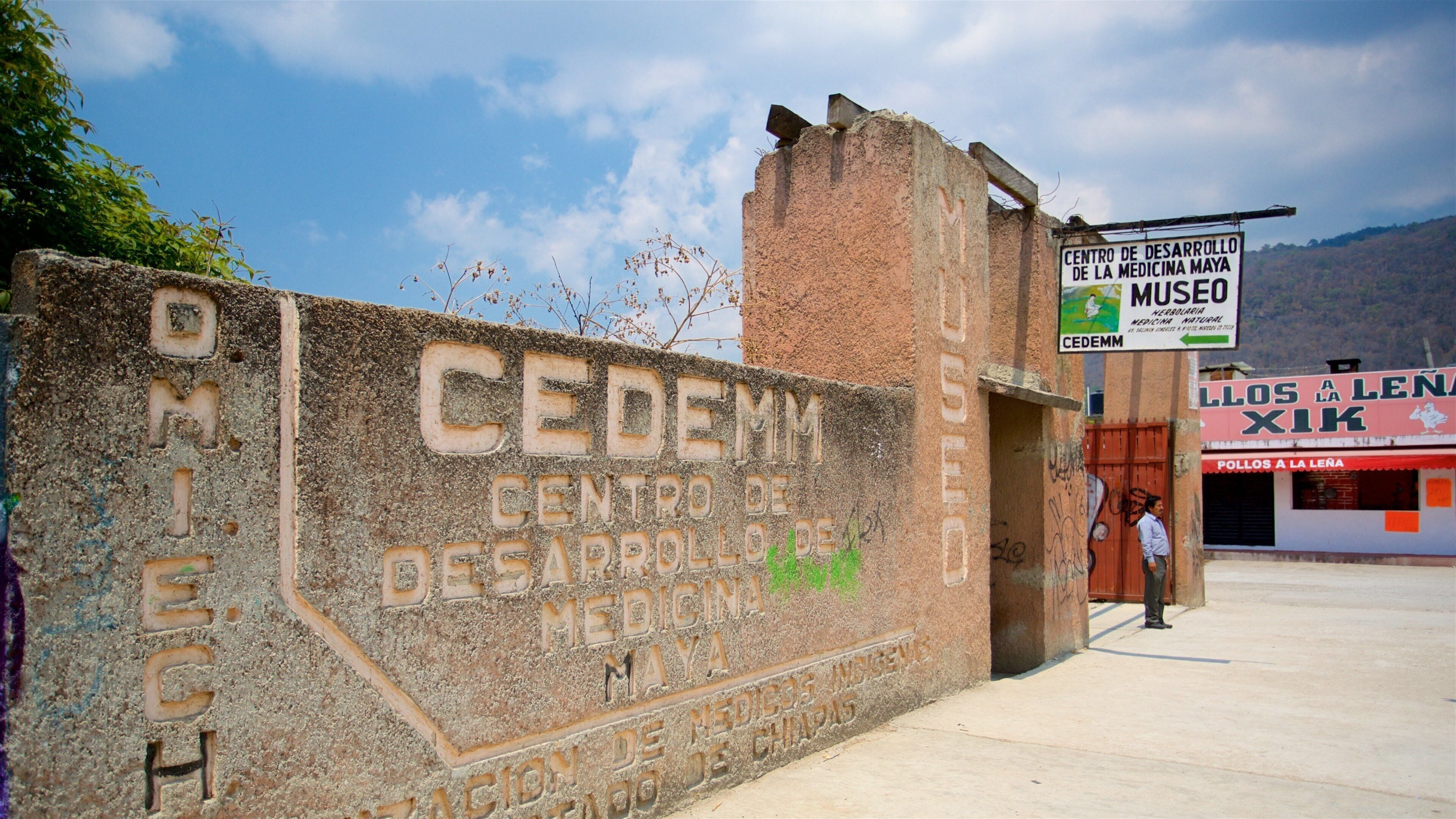 A weathered stone wall with inscriptions, a sign for 'Centro de Desarrollo de Medicina Maya', and a building with a red awning.
