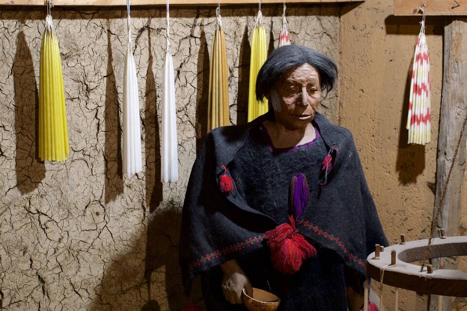 A person in traditional attire holding a bowl, standing in a room with a cracked wall and hanging ceremonial objects.
