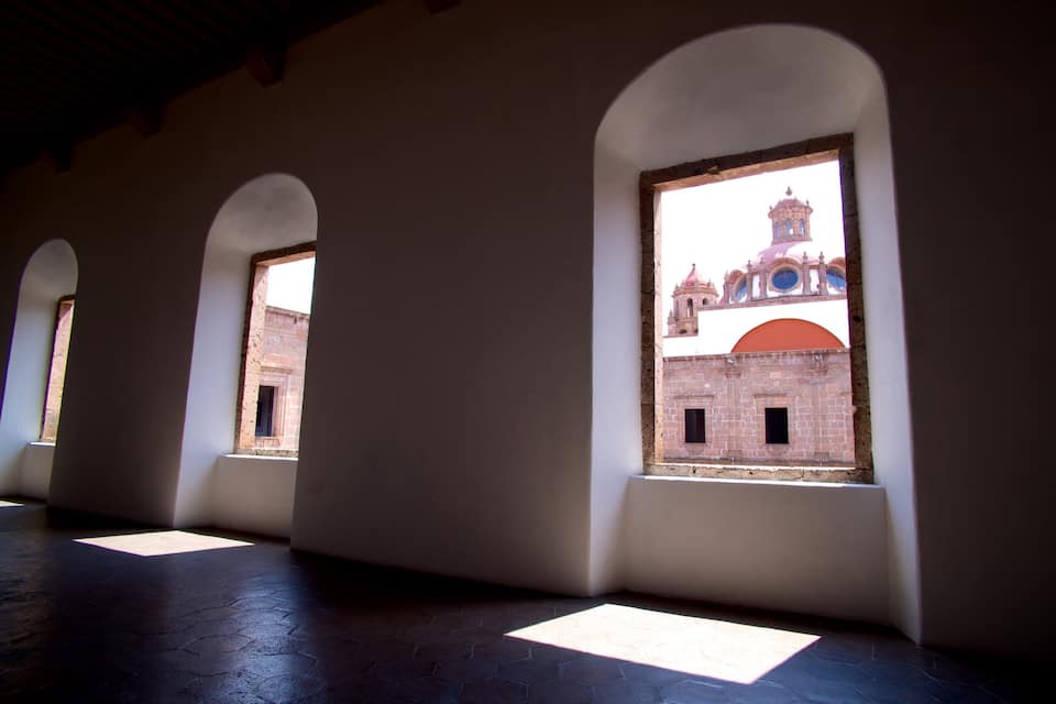 Un pasillo con ventanales arqueados que dan a un edificio histórico con una torre de campanas.