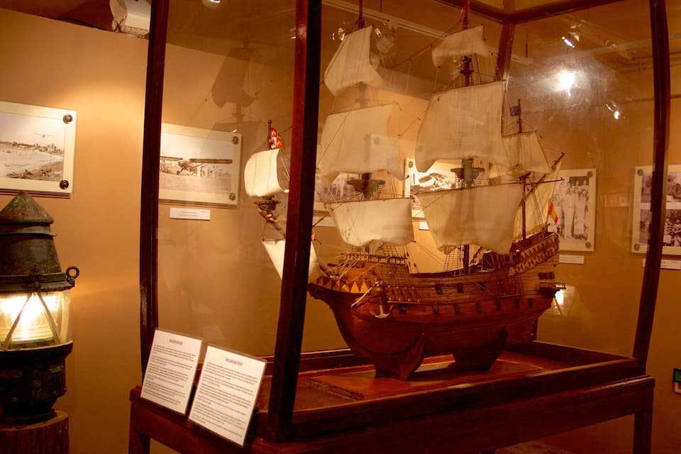 A historical ship model displayed in a glass case, with informational plaques and a vintage lantern beside it.