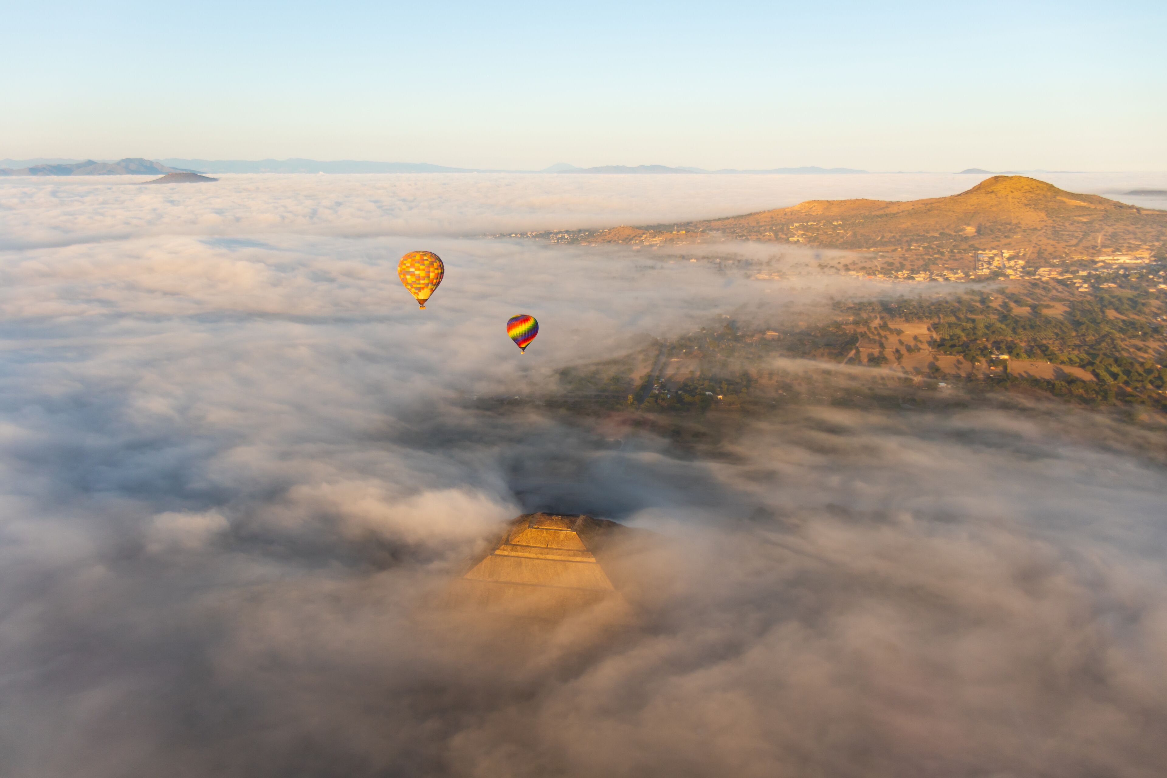 Hot air balloons rising above a dense cloud cover during sunrise, with the golden hues of the morning sun reflecting on the top of the Pyramid of the Sun at Teotihuacan, Mexico