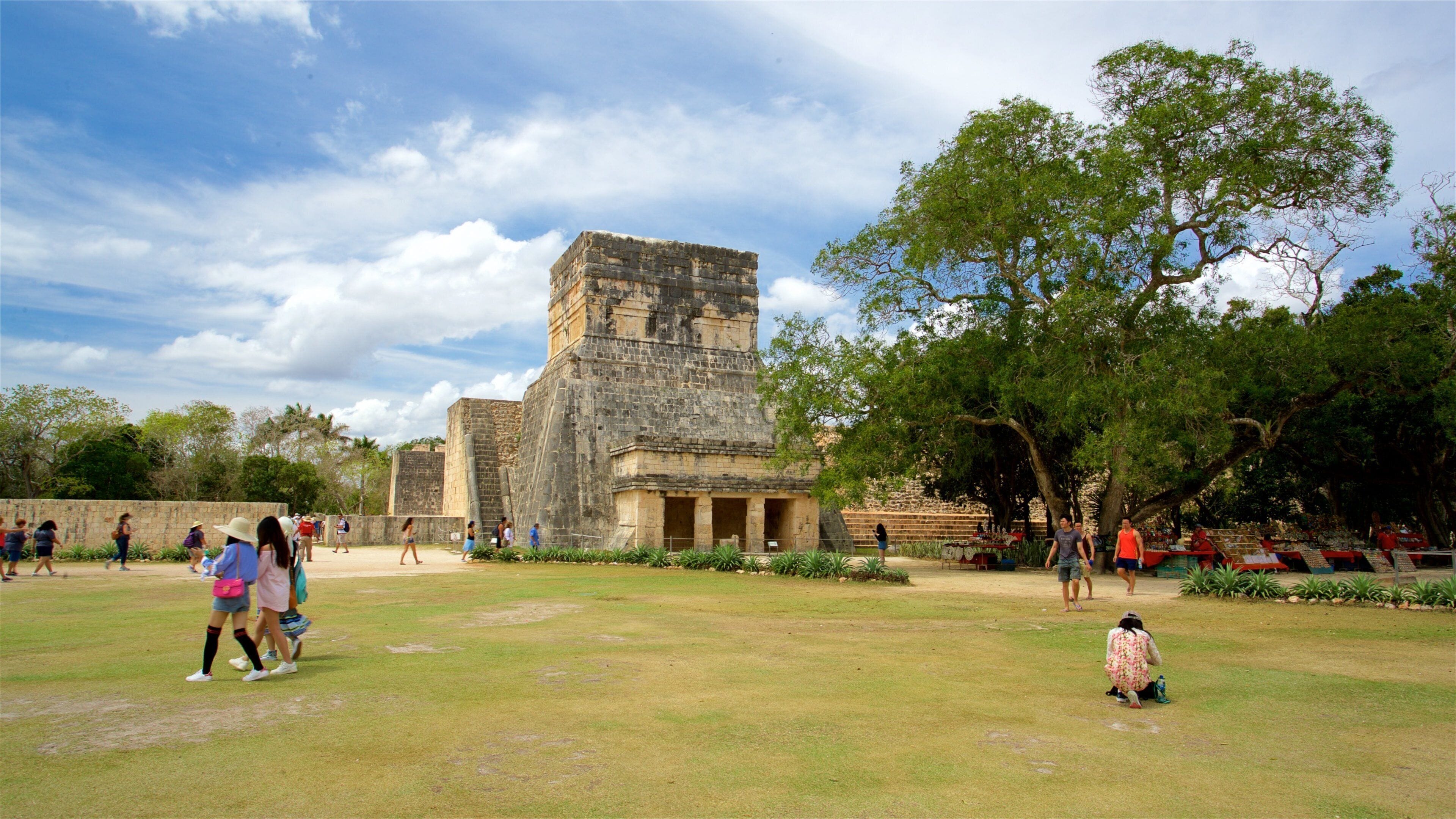 Una gran estructura de piedra antigua rodeada de un área con césped, árboles y gente caminando.