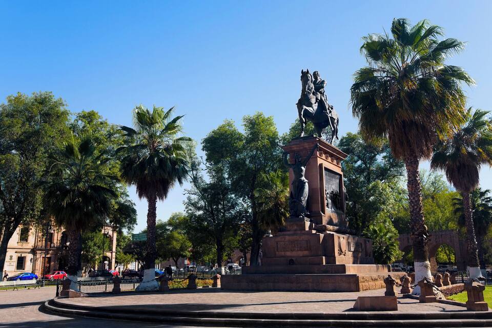 A statue of a man on horseback in a park with palm trees and a building in the background.