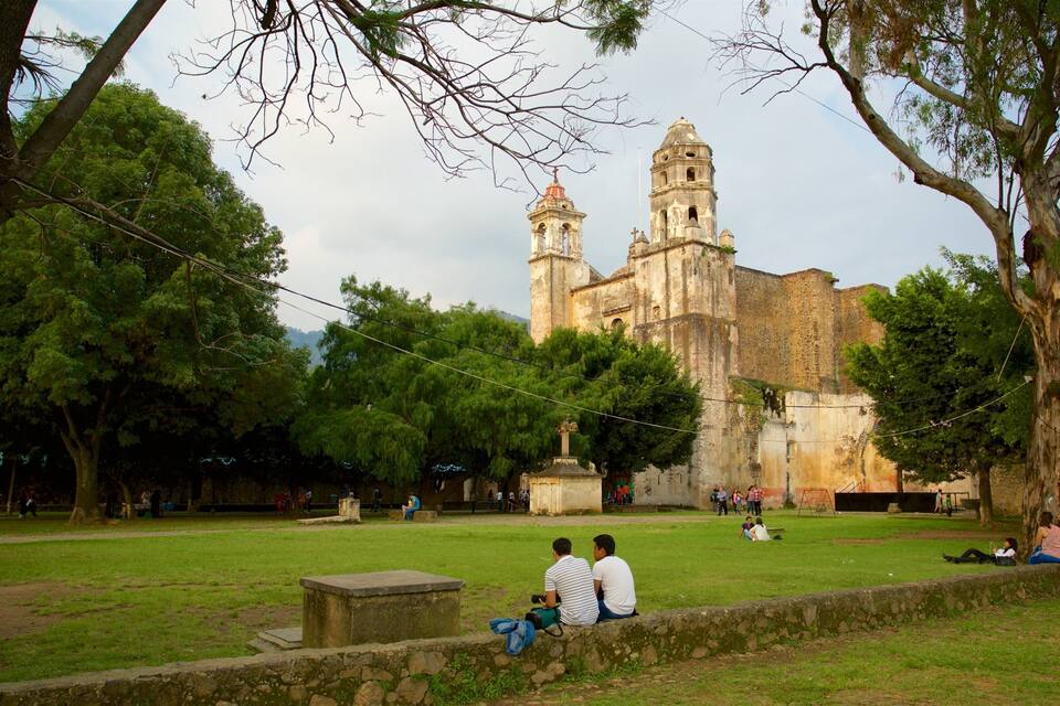 Una iglesia histórica con dos torres, rodeada por un parque arbolado con césped y gente sentada en un muro de piedra.