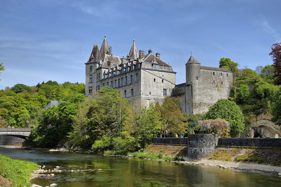 Un château historique doté de nombreuses tours, une rivière au premier plan et une végétation luxuriante qui entoure l’édifice.