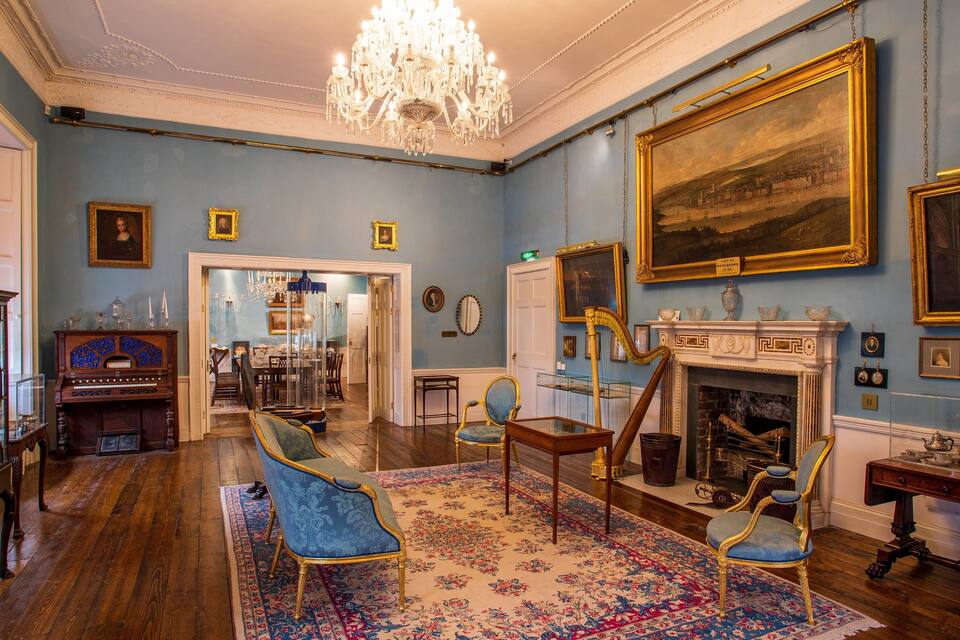 A room with blue walls, wooden floors, a chandelier, a fireplace, and several framed pictures.