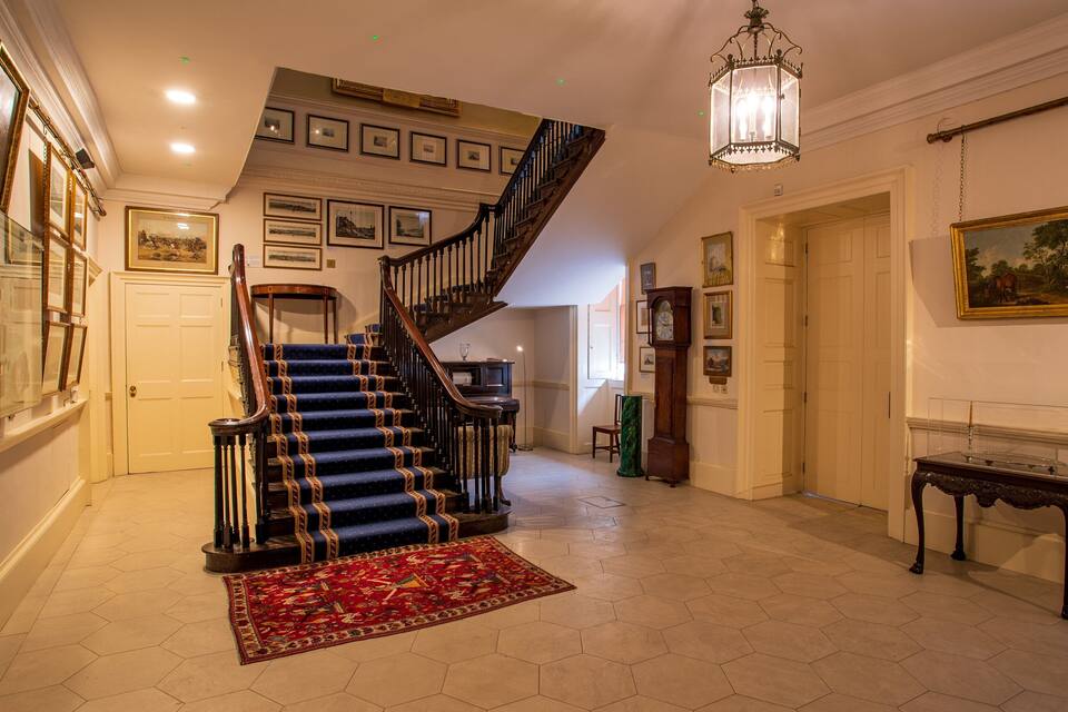 A grand staircase with blue carpet and wooden railings, a chandelier, and framed pictures on the walls.