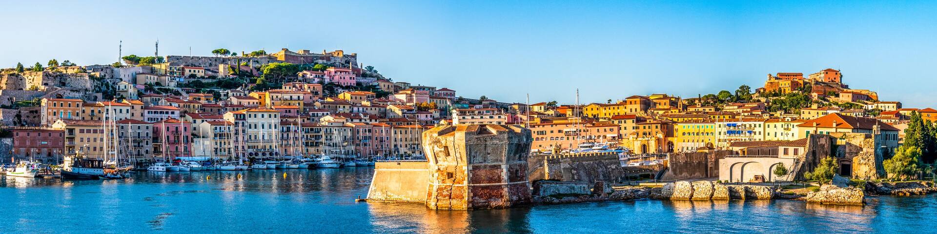 A coastal town with historic buildings, a fortified structure, and a clear blue sea.