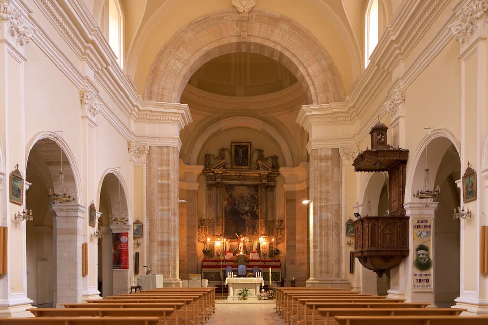 Interior de una iglesia con techos abovedados, bancos de madera y un altar central con estatuas religiosas y velas.