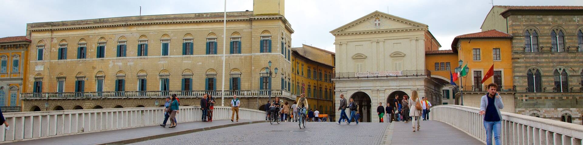 Un puente empedrado con gente caminando y andando en bicicleta, una torre del reloj y edificios históricos de arquitectura clásica.