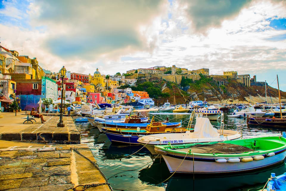 A harbor with colorful boats docked, a historic building on a hill, and a stone-paved waterfront.
