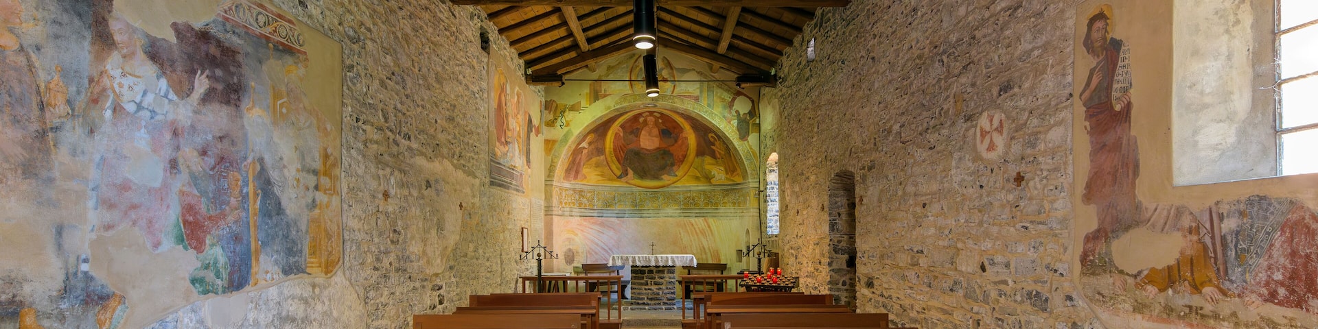 Interior de una iglesia con bancos de madera, paredes de piedra y frescos coloridos.