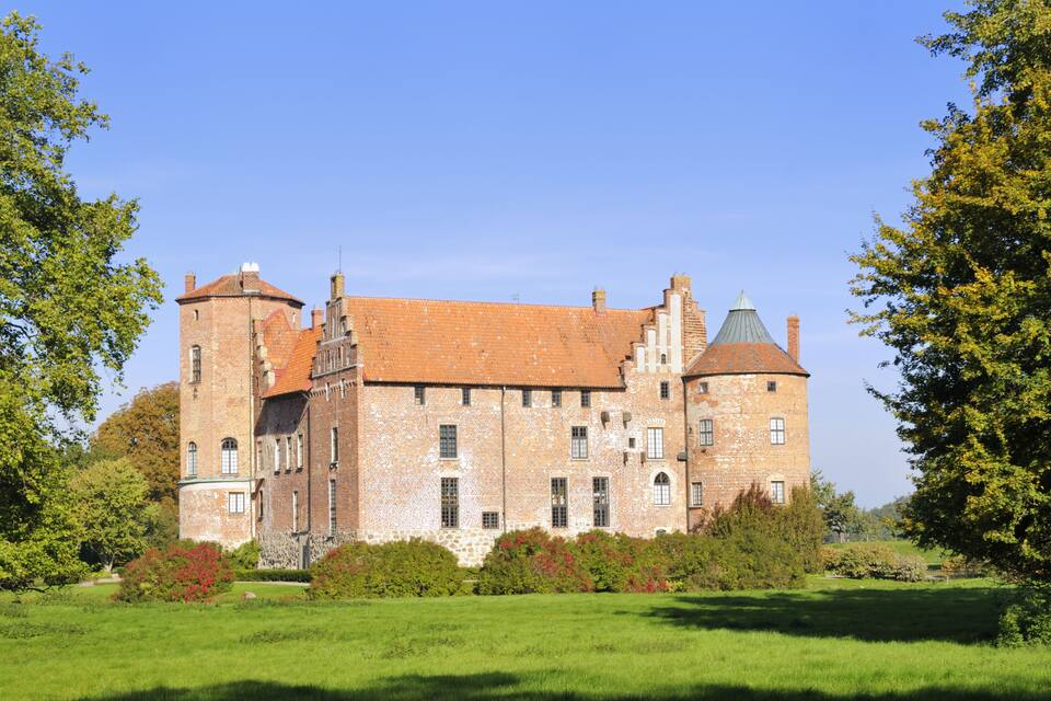 A large, historic brick castle with a red-tiled roof, surrounded by lush green grass and trees.