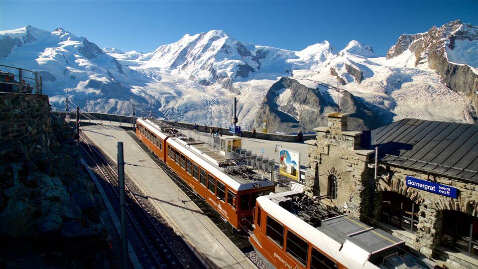 A mountain railway with a train at a station, snow-covered peaks in the background, and a stone building with a sign.