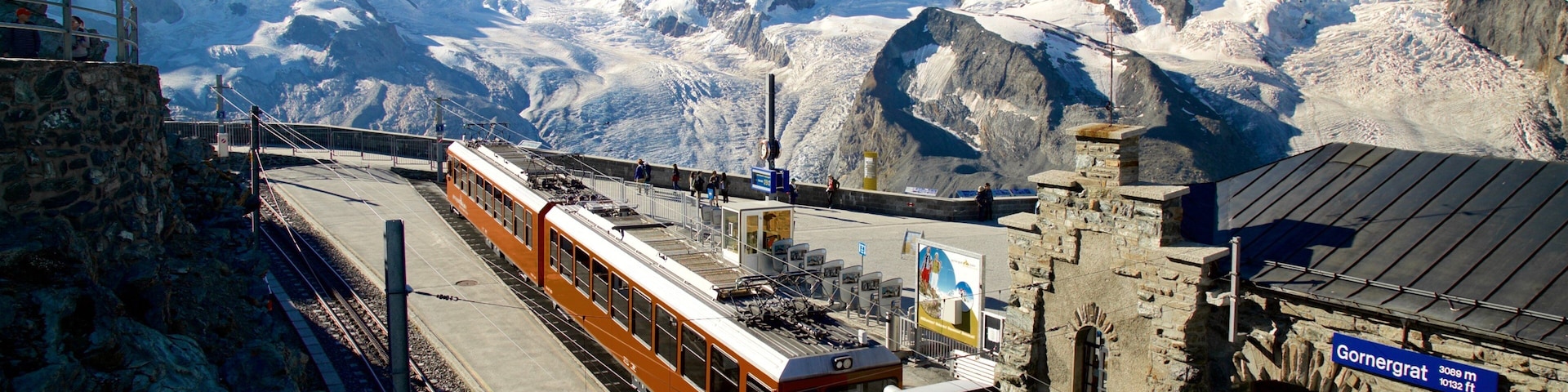 A mountain railway with a train at a station, snow-covered peaks in the background, and a stone building with a sign.