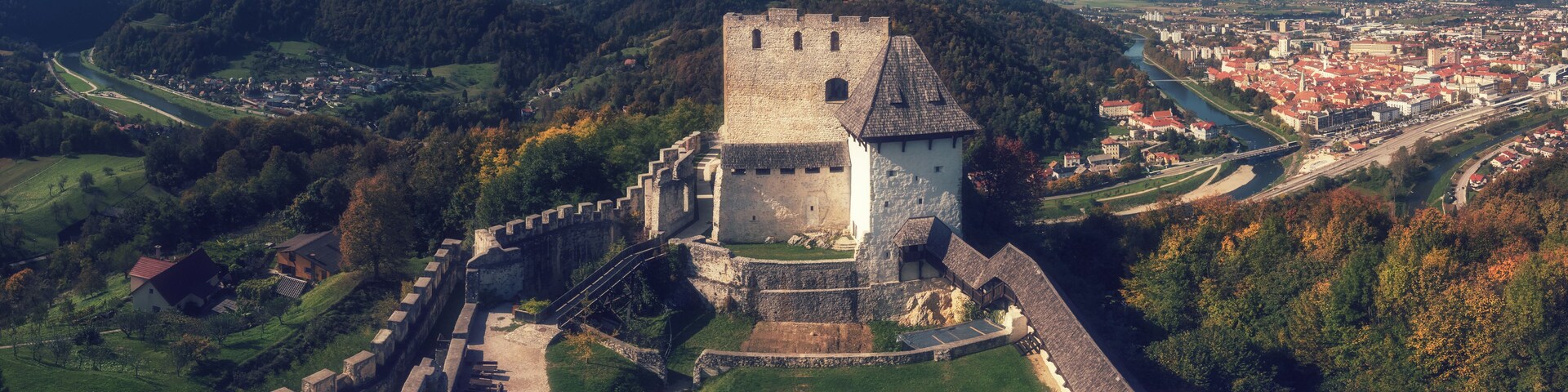 Un castillo histórico con un foso, rodeado de exuberante vegetación y montañas al fondo.