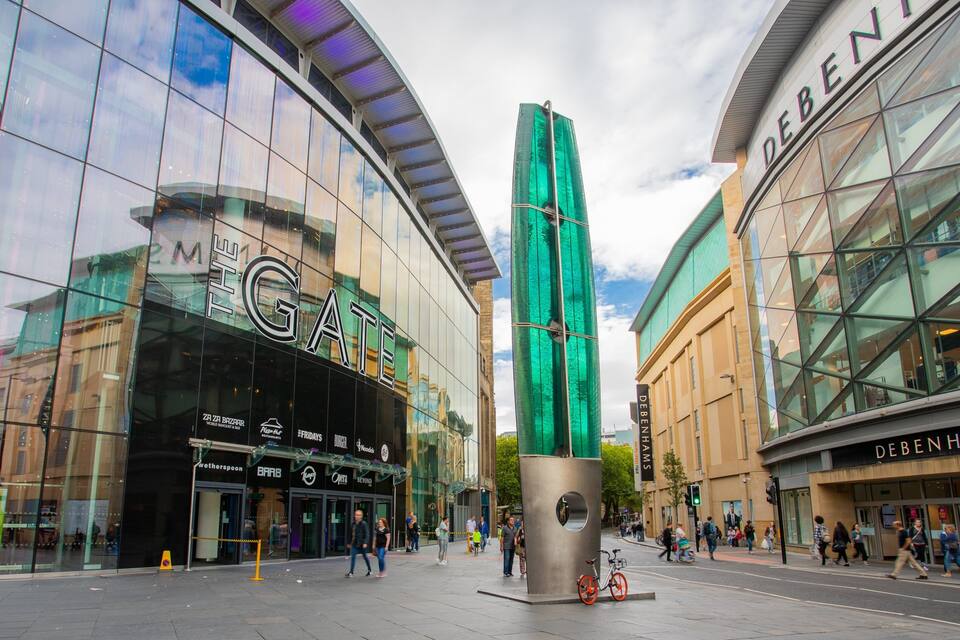 Zona comercial moderna con tiendas de fachada de vidrio, incluyendo 'The Gate' y 'Debenhams'.