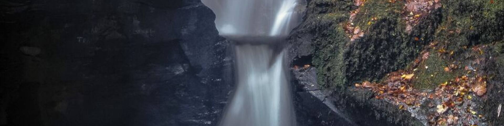 Una cascada desciende entre un profundo desfiladero rocoso, con paredes cubiertas de musgo y un pequeño estanque en la base.