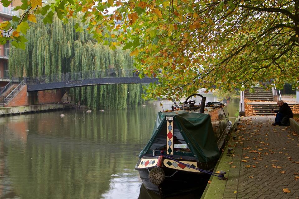 A canal with a narrowboat docked at the edge, a bridge in the background, and a tree with autumn leaves.