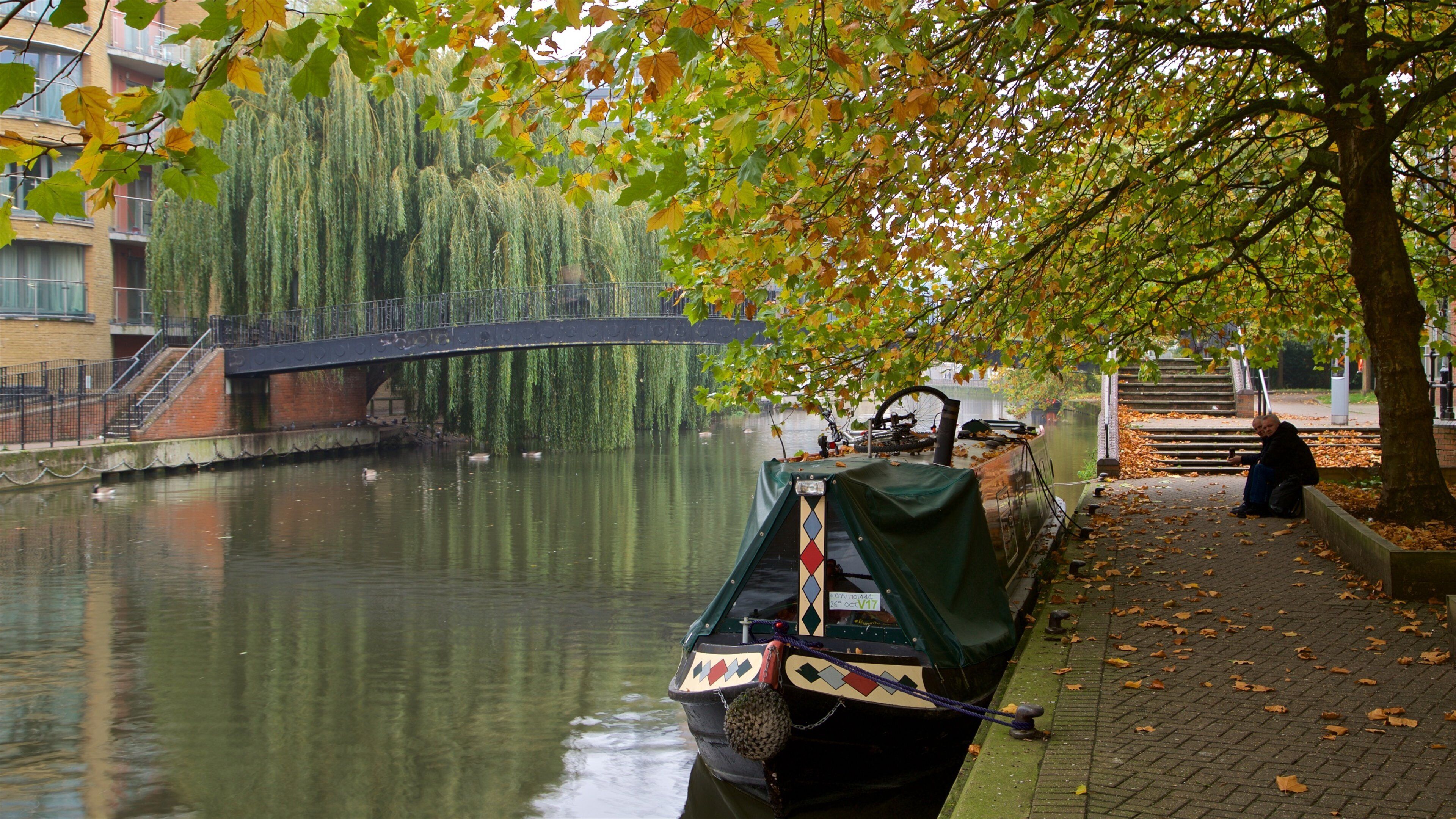 A canal with a narrowboat docked at the edge, a bridge in the background, and a tree with autumn leaves.
