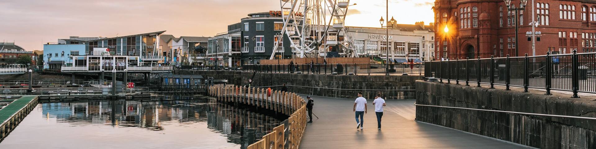 A waterfront promenade with a large Ferris wheel, a historic clock tower, and modern buildings.