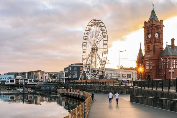 A waterfront promenade with a large Ferris wheel, a historic clock tower, and modern buildings.