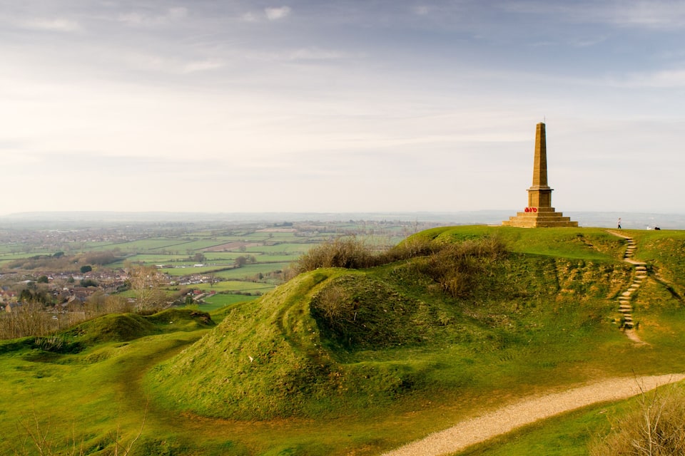 A monument on a grassy hill with a path leading up to it, overlooking a valley with fields and a small town.