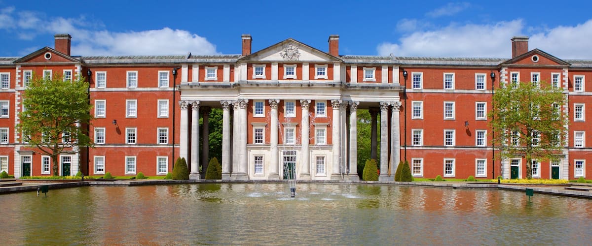 A large red brick building with a central entrance flanked by columns, a fountain in front, and trees on either side.