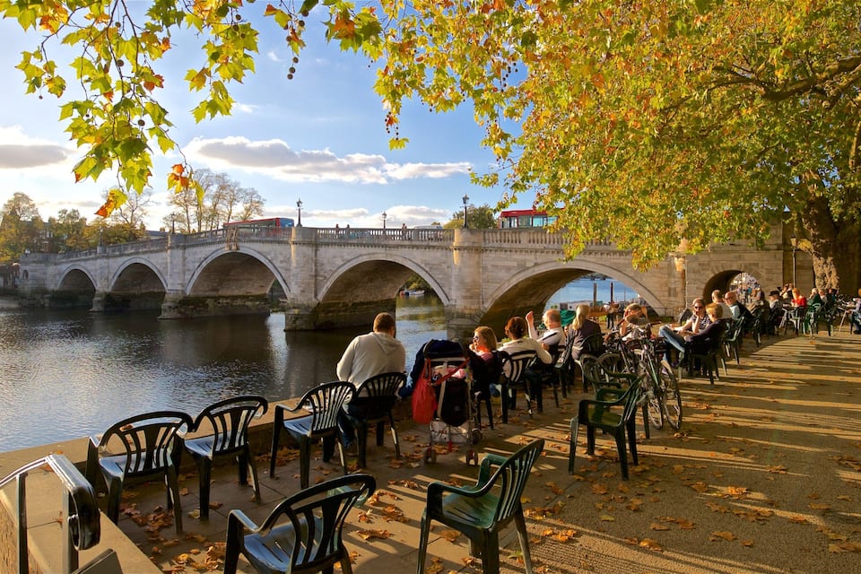 A stone bridge over a river with people sitting on chairs and tables on the riverbank.