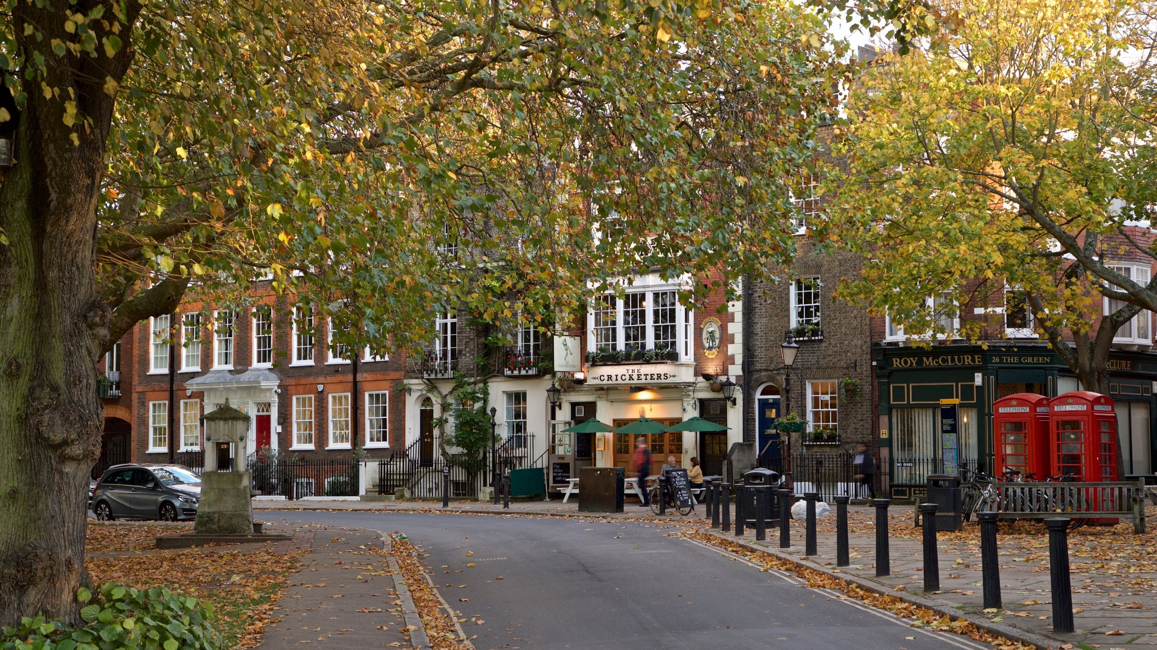 A tree-lined street with buildings, a pub named 'The Cricketers', and red telephone booths.