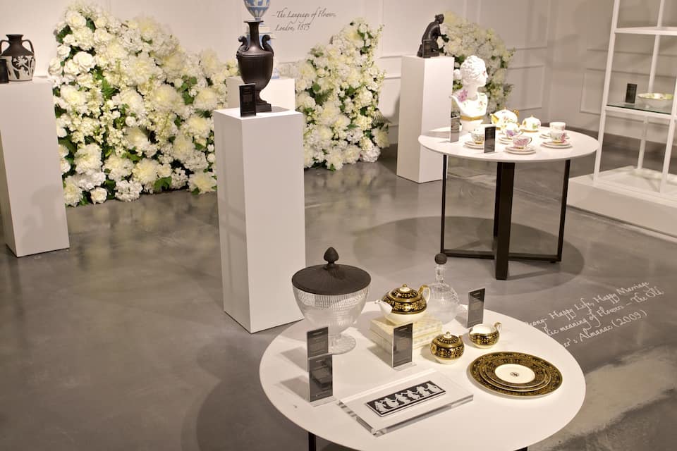 A museum exhibit with white floral arrangements, porcelain tea sets, and decorative vases.