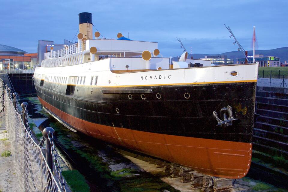 Un gran barco llamado 'Nomadic' atracado en un puerto con un paseo marítimo de piedra y edificios industriales al fondo.