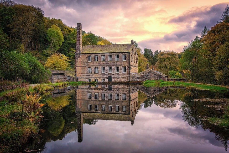 Un edificio histórico de piedra reflejado en un lago tranquilo, rodeado de follaje otoñal.